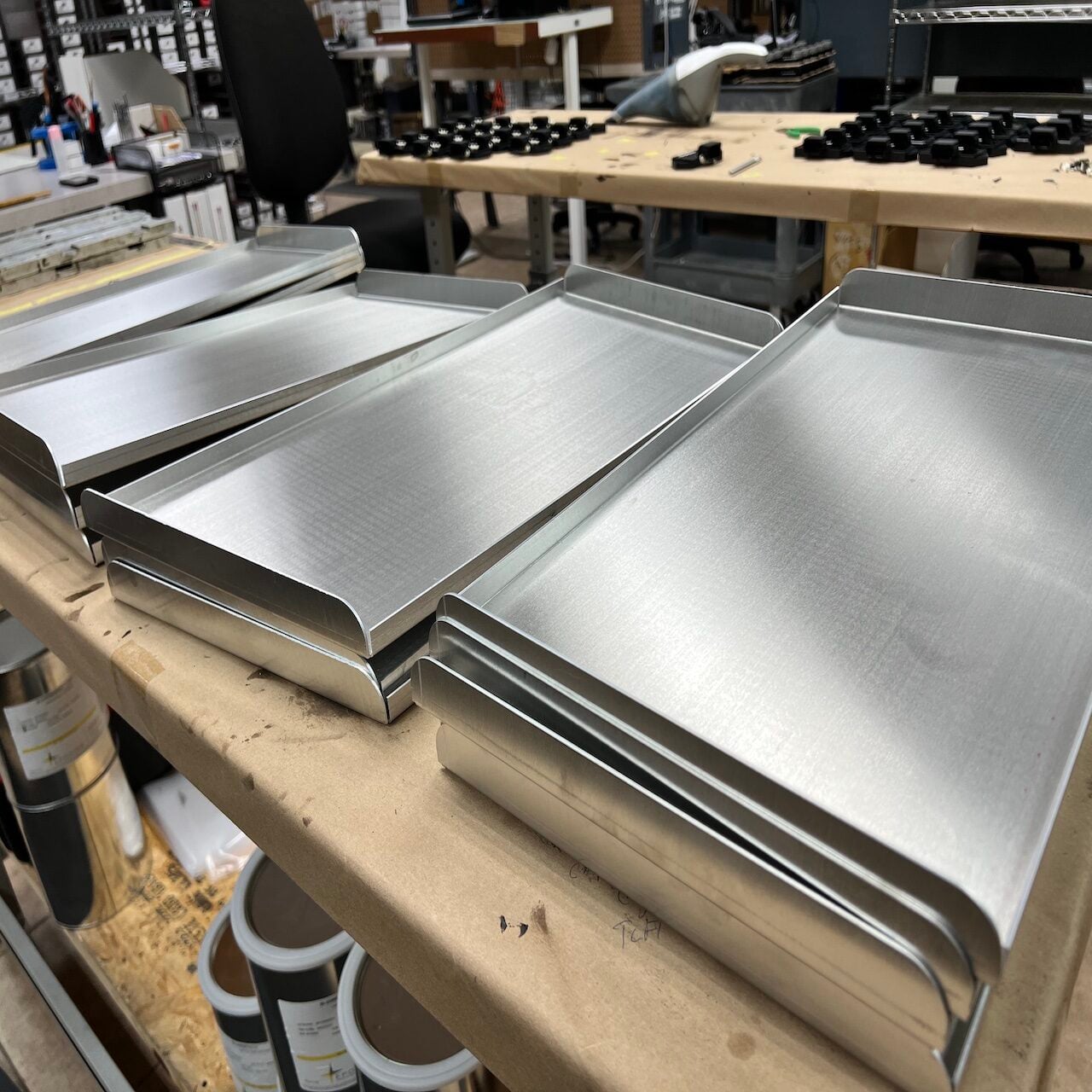 Several rectangular metal trays with raised edges are stacked on a workbench in a workshop, with tools and materials visible in the background.