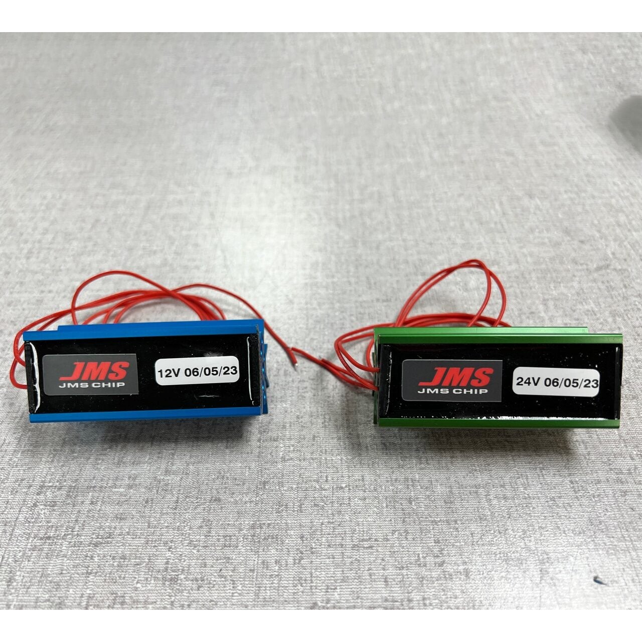 Two electronic modules labeled JMS CHIP sit side by side on a gray surface. The left module is blue and labeled 12V 06/05/23, and the right module is green and labeled 24V 06/05/23. Both have red wires attached.
