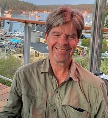 A smiling older man with gray hair sits outdoors on a patio overlooking a marina with boats, water, and trees in the background. He is wearing a light green button-up shirt.