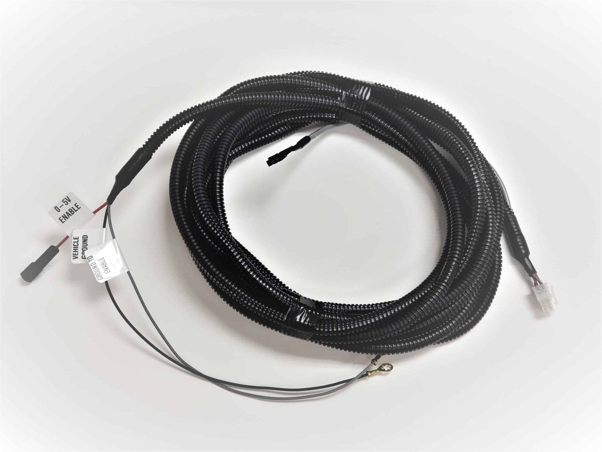 A coiled black electrical wiring harness with a protective sleeve, labeled wires, connectors, and a metal ring terminal, placed on a white surface.