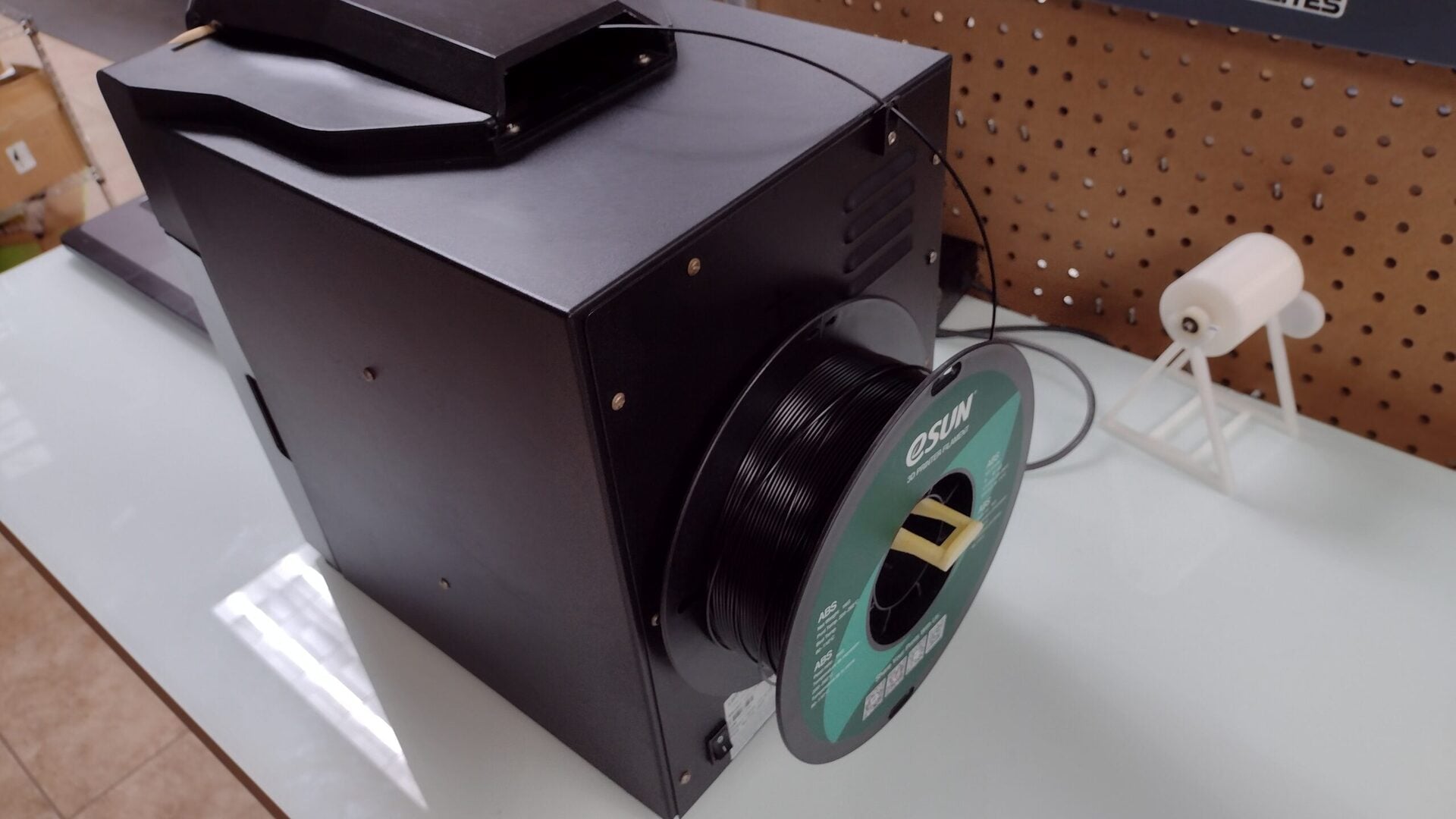 Black 3D printer filament dryer with a spool of black filament loaded, sitting on a white desk near a pegboard wall and a connected power cable.