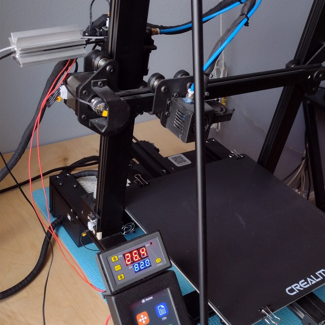 A close-up of a Creality 3D printer on a desk, showing its control panel with a digital display reading 28.9, wires, and the print bed ready for use.