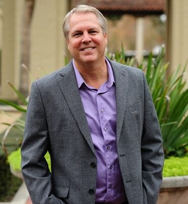 A man with short, light hair wearing a gray blazer and a purple shirt stands outdoors in front of green plants and a building, smiling at the camera.