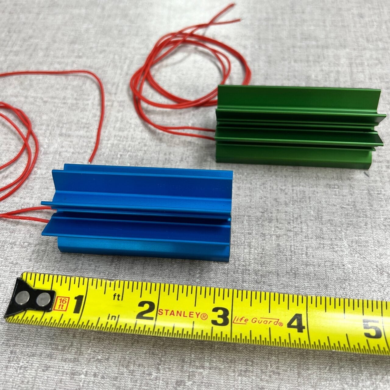 Two colored metal objects, one blue and one green, with red wires attached, rest on a gray surface near a yellow measuring tape showing inches for scale.