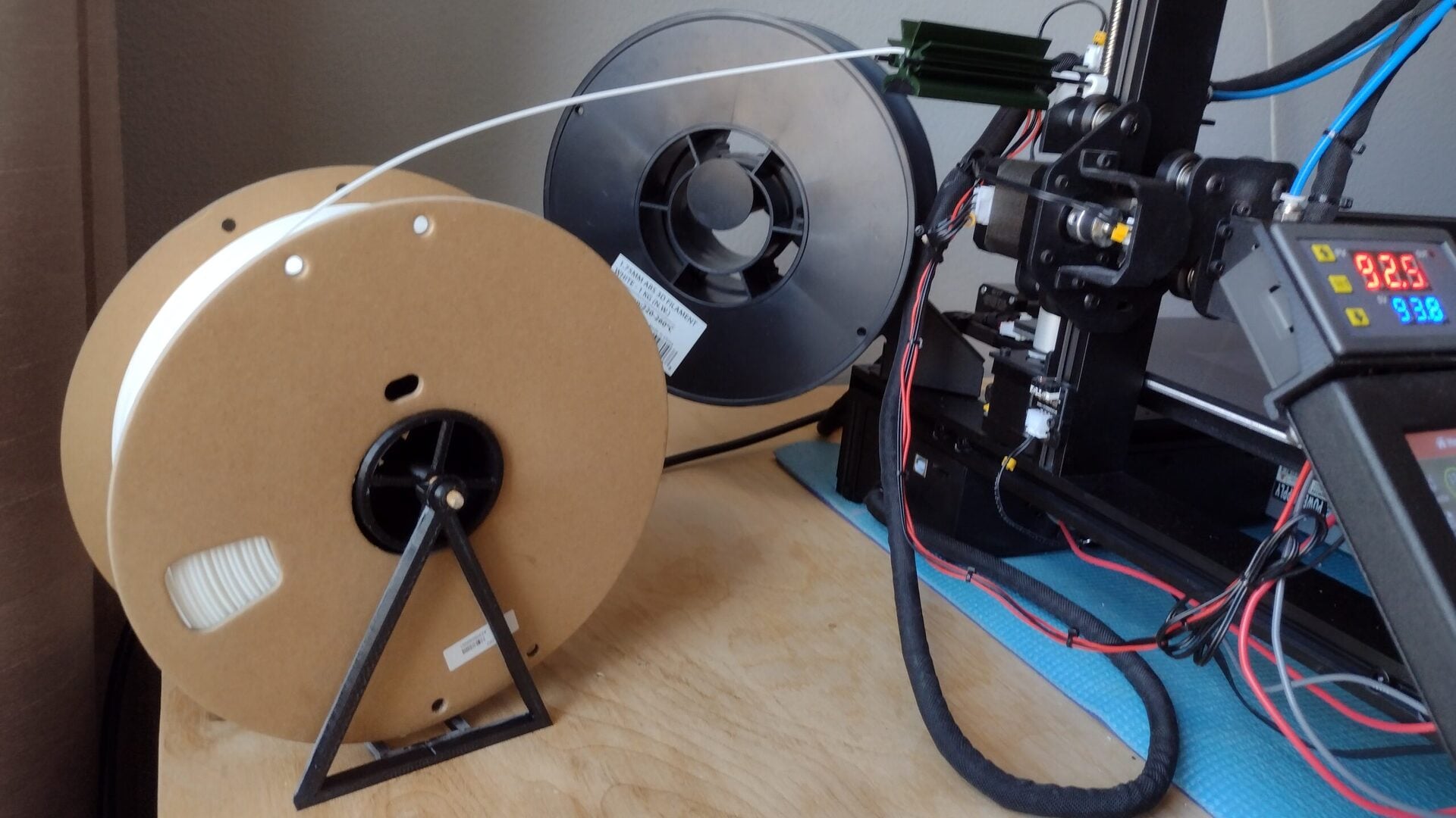 A close-up of a 3D printer setup on a wooden surface, showing two spools of filament, one on a stand feeding filament into the printer, with the printer’s control panel displaying numbers.
