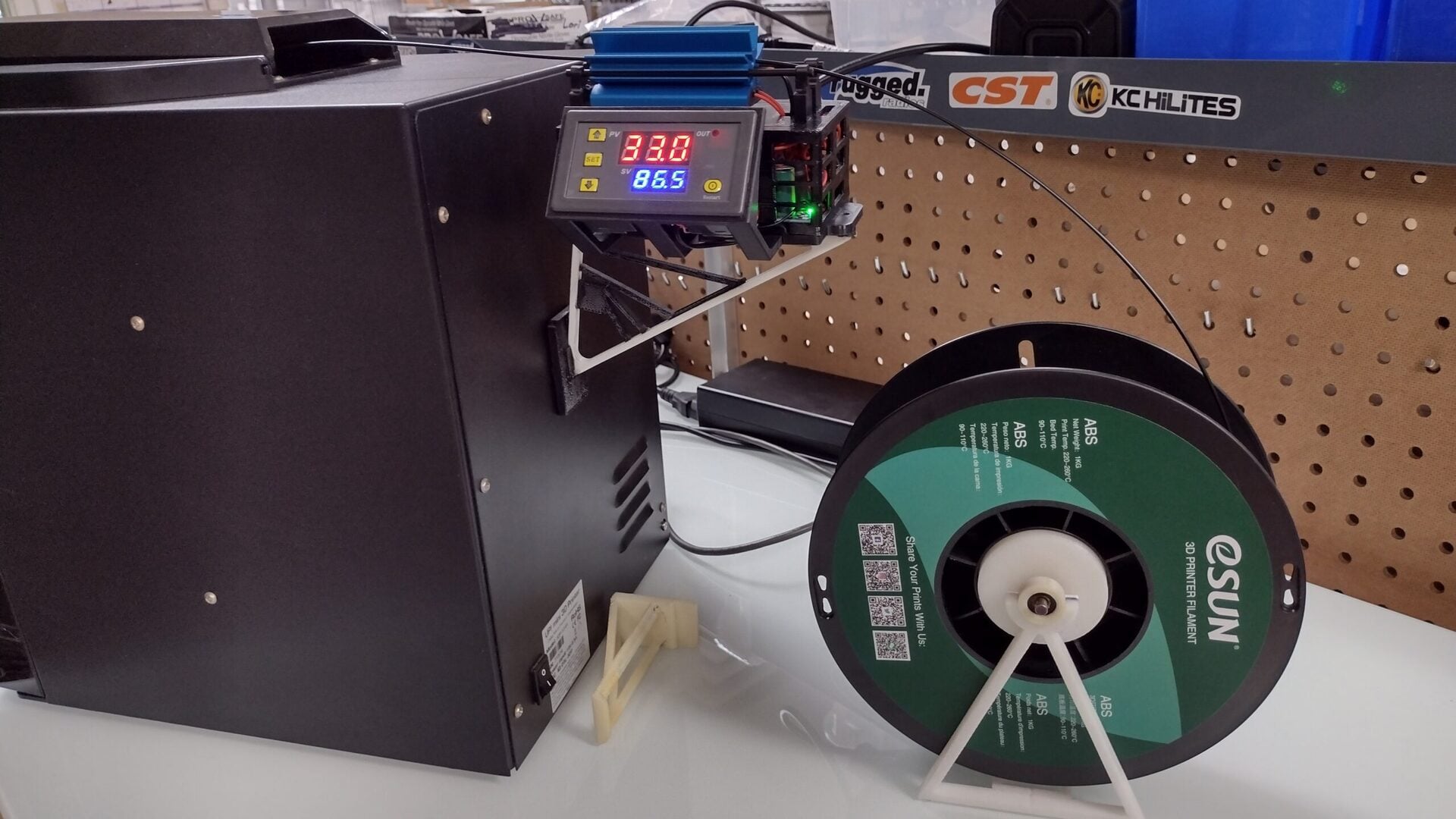 A black filament dryer with a digital display shows temperature and humidity. Next to it, a green spool of eSUN 3D printing filament rests on a white stand. The setup is on a workbench with pegboard and stickers in the background.