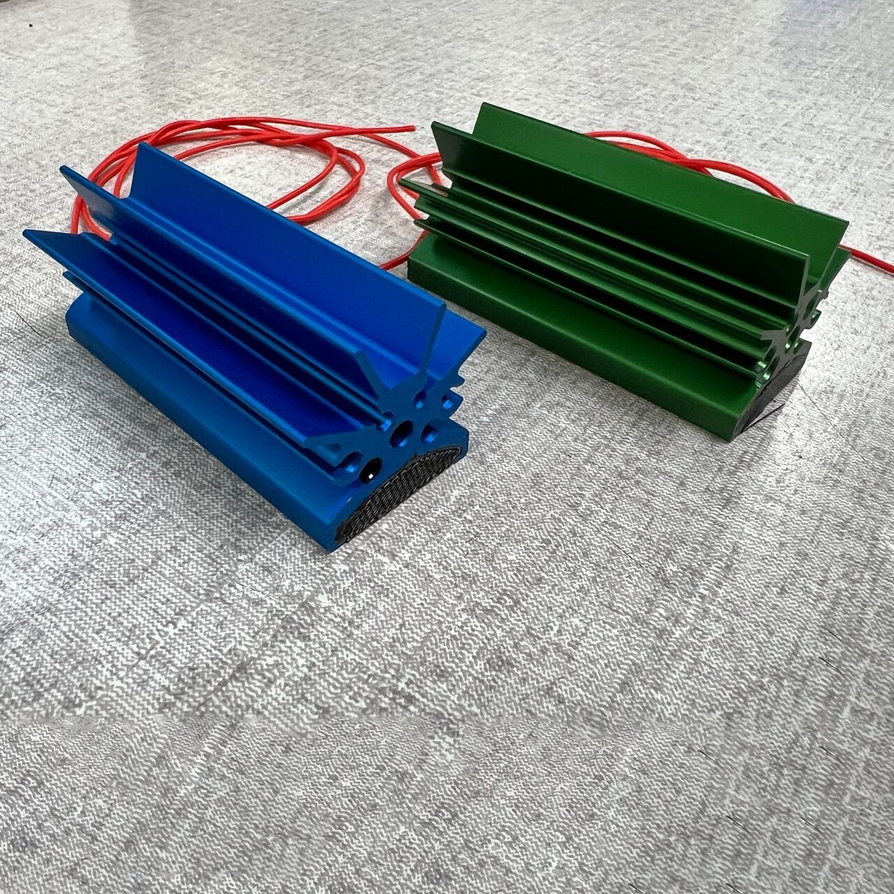 Two rectangular electronic devices with large heat sinks, one blue and one green, each with red wires attached, are placed on a textured gray surface.