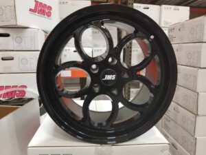 A glossy black alloy wheel with a five-spoke design is displayed upright. The center cap features the JMS logo. Stacks of white boxes, some labeled with JMS, are seen in the background.
